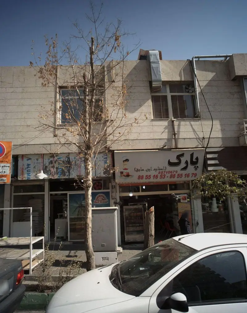 تصویر نمای بیرونی فست فود پارک، با تابلوی مشخص، ورودی روشن، یک درخت پاییزی و ماشین سفید در جلوی مغازه.