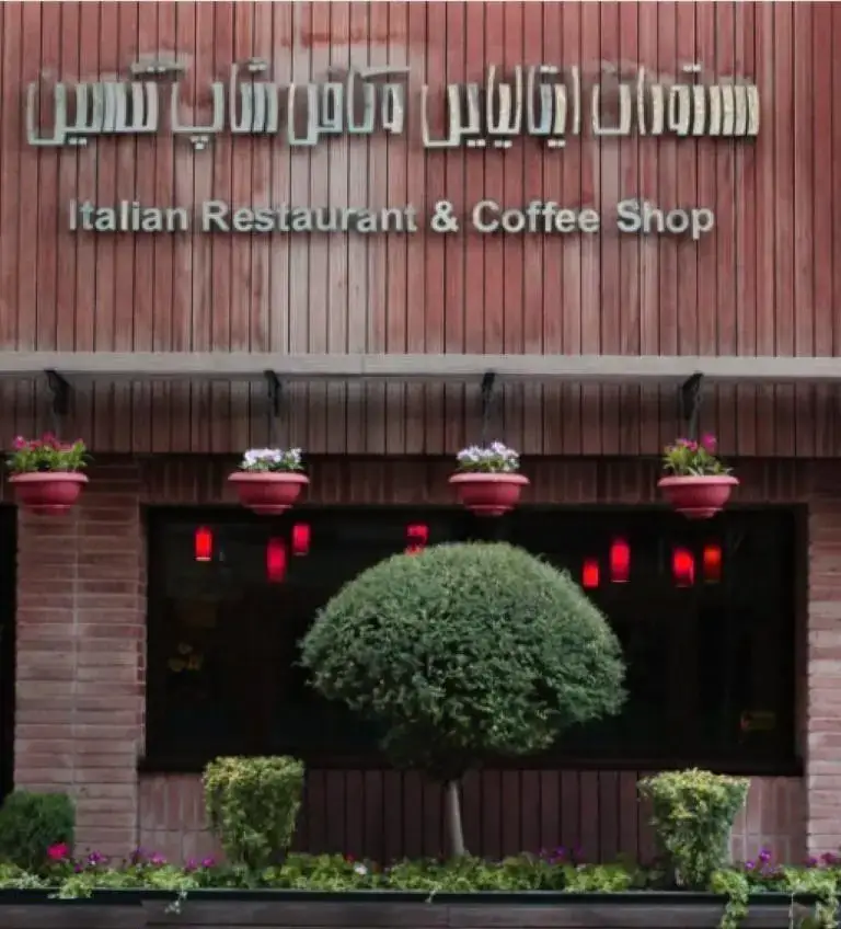 نمای بیرونی رستوران و کافی شاپ ایتالیایی تکسین با طراحی چوبی و آجر، گلدان‌های آویز گل و درختچه زیبا در ورودی.