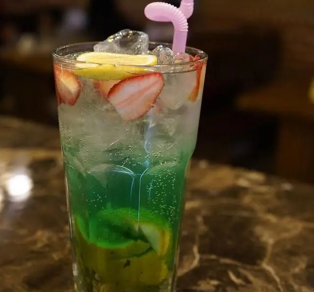 یک نوشیدنی خنک و گازدار میوه‌ای با لایه‌های سبز نعنایی و شفاف با توت فرنگی، لیمو و یخ در کافه رستوران دورهمی امامزاده حسن.