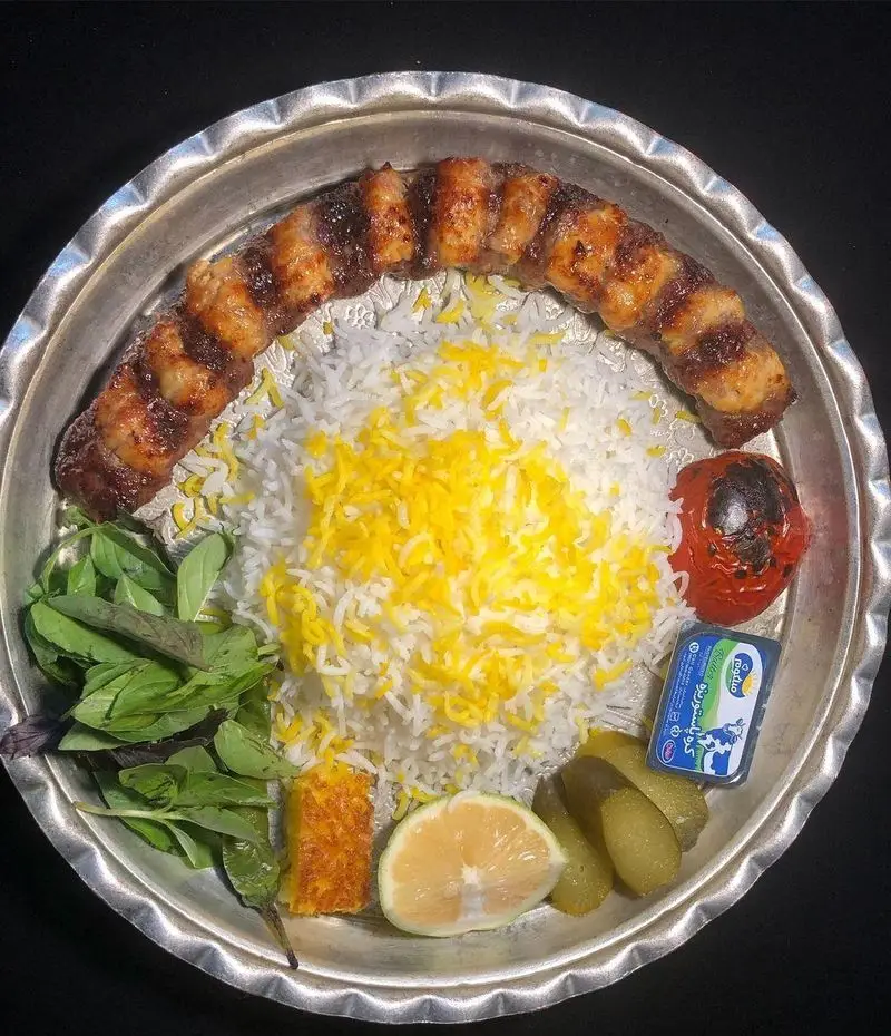 سرو چلوکباب لذیذ ایرانی شامل کباب، برنج زعفرانی، گوجه کبابی، سبزی تازه، لیمو و خیارشور در کافه رستوران دورهمی امامزاده حسن.