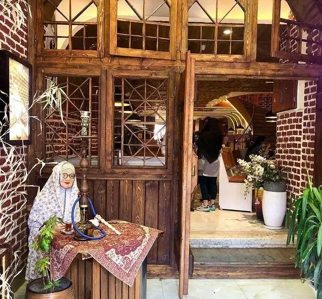 نمای ورودی کافه رستوران دورهمی امامزاده حسن با دکوراسیون چوبی، پنجره‌های سنتی و آدمک پیرزن قلیان‌به‌دست.