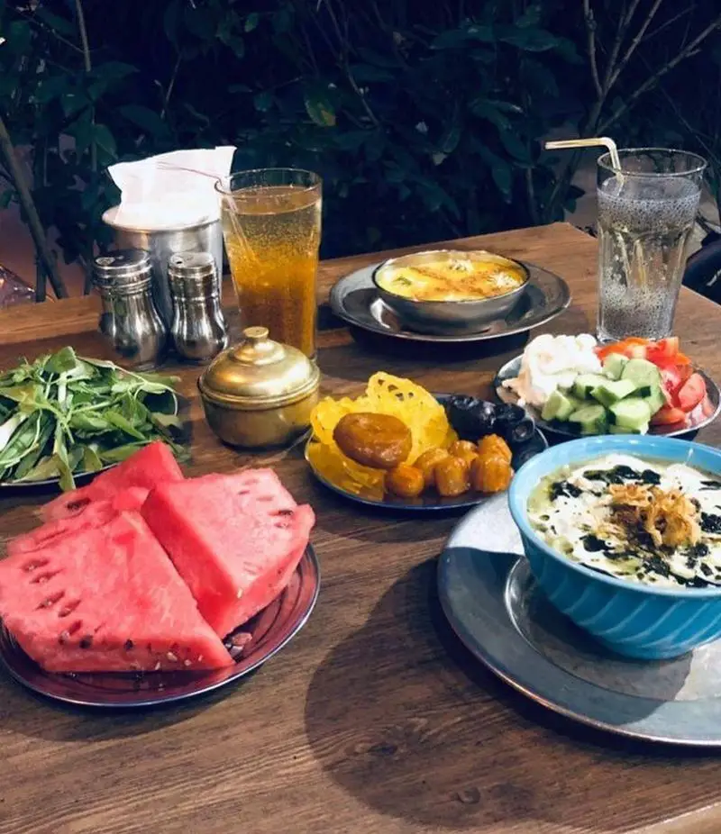 سفره رنگین افطار کافه رستوران عمو سهراب: آش، هندوانه، سبزی، خرما، زولبیا بامیه، سالاد و نوشیدنی‌های خنک در فضایی دلپذیر.