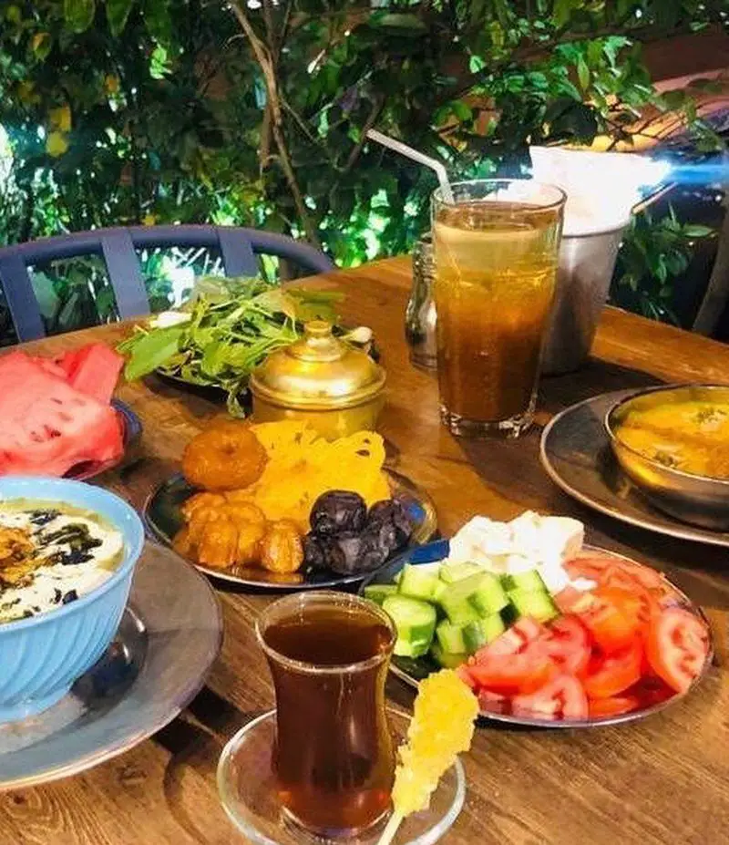 میز صبحانه سنتی و کامل کافه رستوران عمو سهراب: آش، چای، زولبیا بامیه، سبزی خوردن، پنیر و میوه در فضایی باغی.