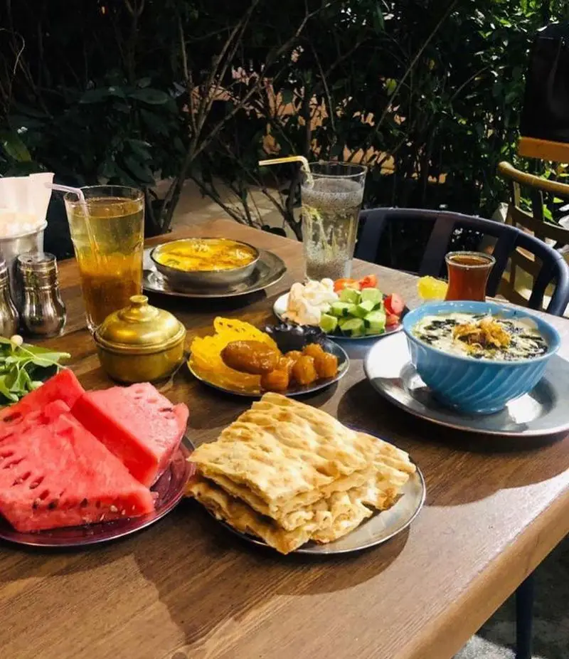 میز چوبی پر از غذاهای سنتی و نوشیدنی‌های ایرانی در کافه رستوران عمو سهراب، شامل آش، نان، هندوانه، شیرینی و سالاد در فضای باز دلنشین.