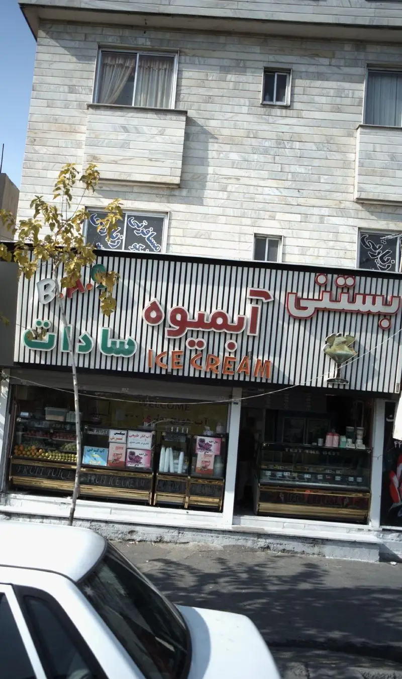 تصویر نمای خارجی مغازه بستنی و آبمیوه سادات با تابلوی چشمگیر، ویترین‌هایی مملو از میوه‌های تازه، آبمیوه‌ها و انواع بستنی.