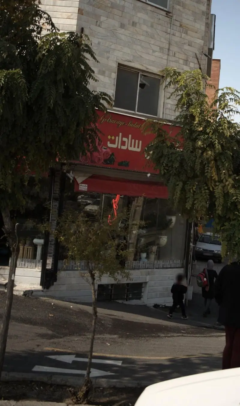 نمای بیرونی مغازه بستنی و آبمیوه سادات با تابلوی قرمز رنگ، زیر سایه درختان سبز. ساختمان سنگی و خیابانی شلوغ با مردم و ماشین‌ها.