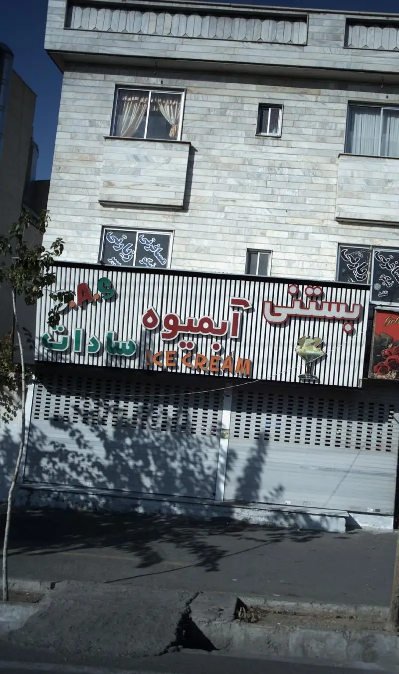 تصویر نمای بیرونی فروشگاه بستنی و آبمیوه سادات در یک ساختمان با نمای سنگی روشن و کرکره‌های پایین، شامل تابلوی فارسی و انگلیسی.