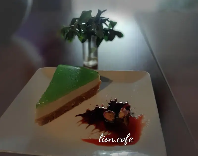 لذت یک دسر شیرین: برش کیک ژله‌ای سبز رنگ با لایه کرمی سفید و بیسکویت، تزئین سس قرمز در کافه لیون.