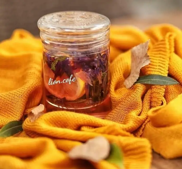 دمنوش گرم پاییزی لیون کافه، ترکیبی از لیمو و گل‌های بنفش در شیشه، روی پس‌زمینه بافتنی زرد. تجربه‌ای دلنشین و آرامش‌بخش از lion.cafe.