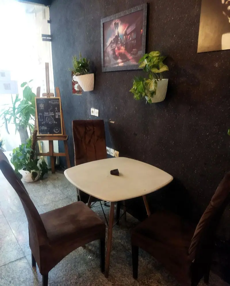 فضای داخلی لیون کافه: دکوراسیون دنج با میز و صندلی شیک، گلدان‌های آویز دیواری، تابلو هنری و تخته سیاه منوی قهوه.
