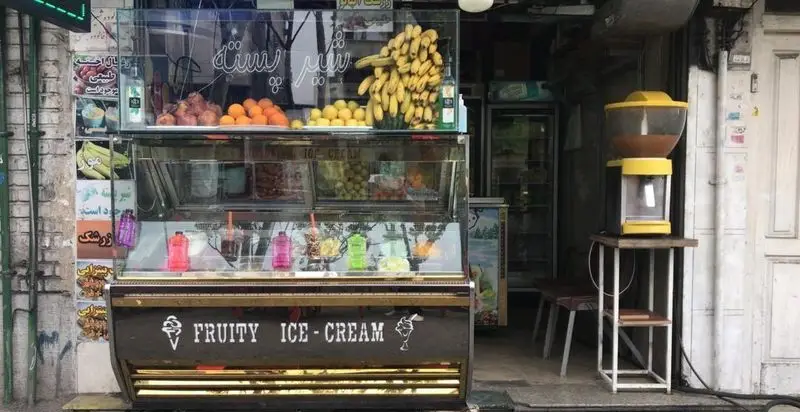 تصویری از مغازه آبمیوه بستنی دلتا: پیشخوان با بستنی های رنگارنگ، میوه های تازه شامل موز، پرتقال و لیمو، و دستگاه آبمیوه. مکانی عالی برای دسر.