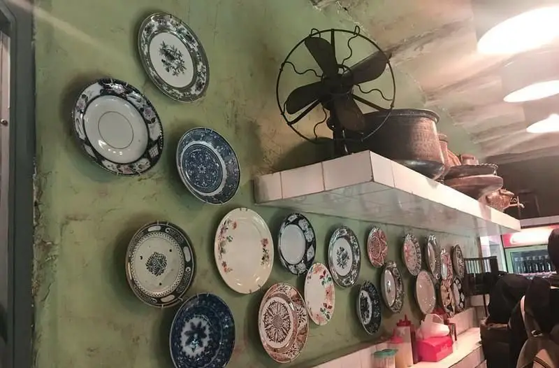 نمایی از دکوراسیون داخلی زیبای رستوران ایرانی آقا خان، شامل بشقاب‌های طرح‌دار سنتی آویزان بر دیوار سبز، پنکه قدیمی و ظروف سفالی.