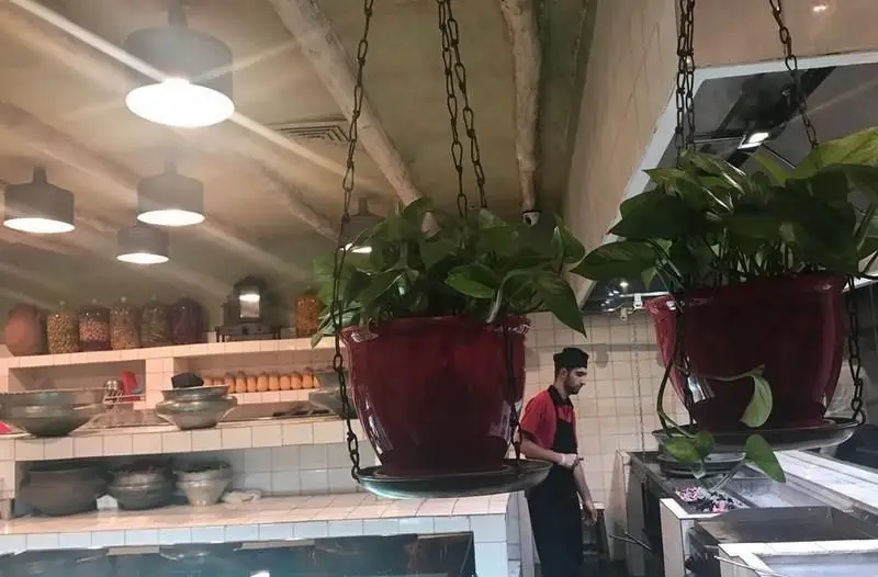 فضای داخلی دلنشین و دکوراسیون سنتی آشپزخانه رستوران ایرانی آقا خان با گلدان‌های آویز و آشپز در حال آماده‌سازی غذاهای اصیل.