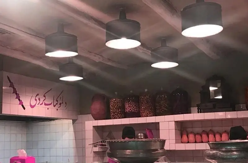 نمای داخلی رستوران ایرانی آقا خان با نورپردازی گرم، دیوارهای کاشی‌کاری شده و قفسه‌های پر از مواد اولیه برای کباب کردی.