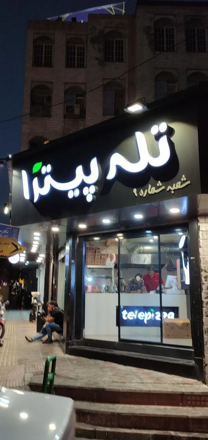 نمای بیرونی شبانگاهی از فست فود تله پیتزا شعبه پاسداران، همراه با تابلوی نئونی روشن و مشتریانی که در حال رفت و آمد هستند.
