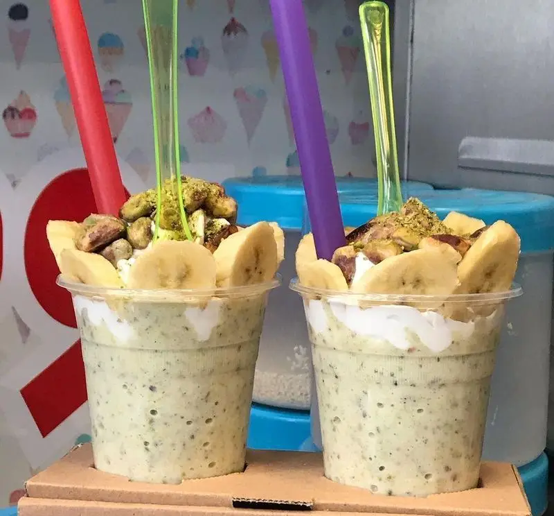 نوشیدنی اسموتی سبز رنگ ابمیوه فردین، تزئین شده با موز، پسته و آجیل، همراه با خامه، یک انتخاب عالی برای روزهای گرم.