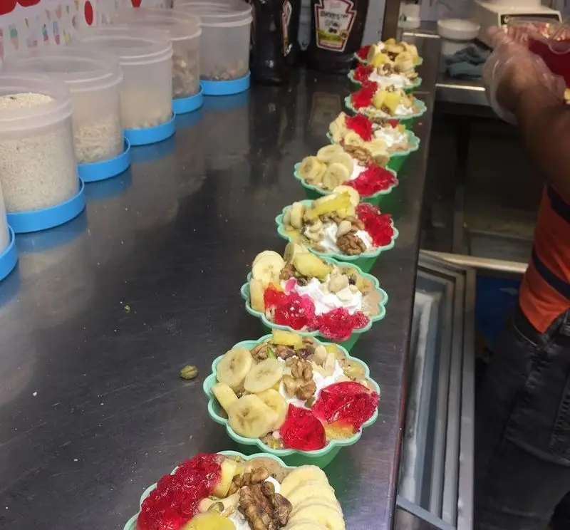 نمایی از دسرهای میوه ای و مغزدار خوشمزه و تازه ابمیوه فردین، شامل موز، خامه و ژله در ظروف سبز آماده سرو برای مشتریان