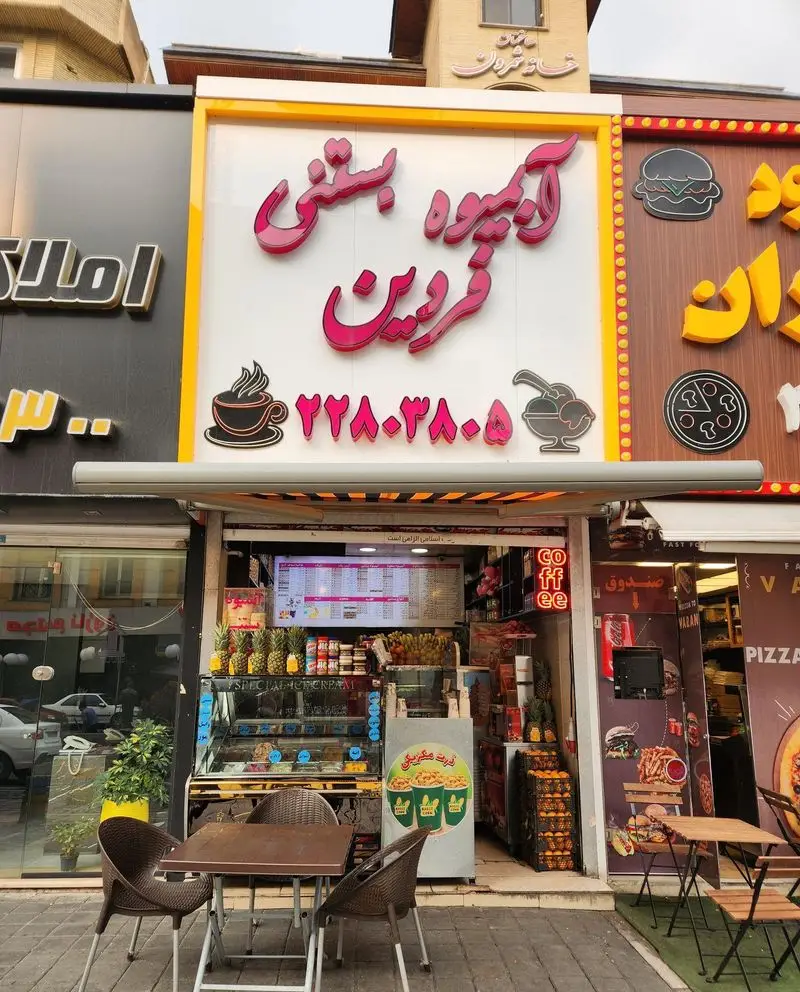 نمای فروشگاه آبمیوه و بستنی فردین با تابلو صورتی برجسته، ویترین پر از میوه‌های تازه، منوی دیجیتال و میز و صندلی در فضای بیرونی.