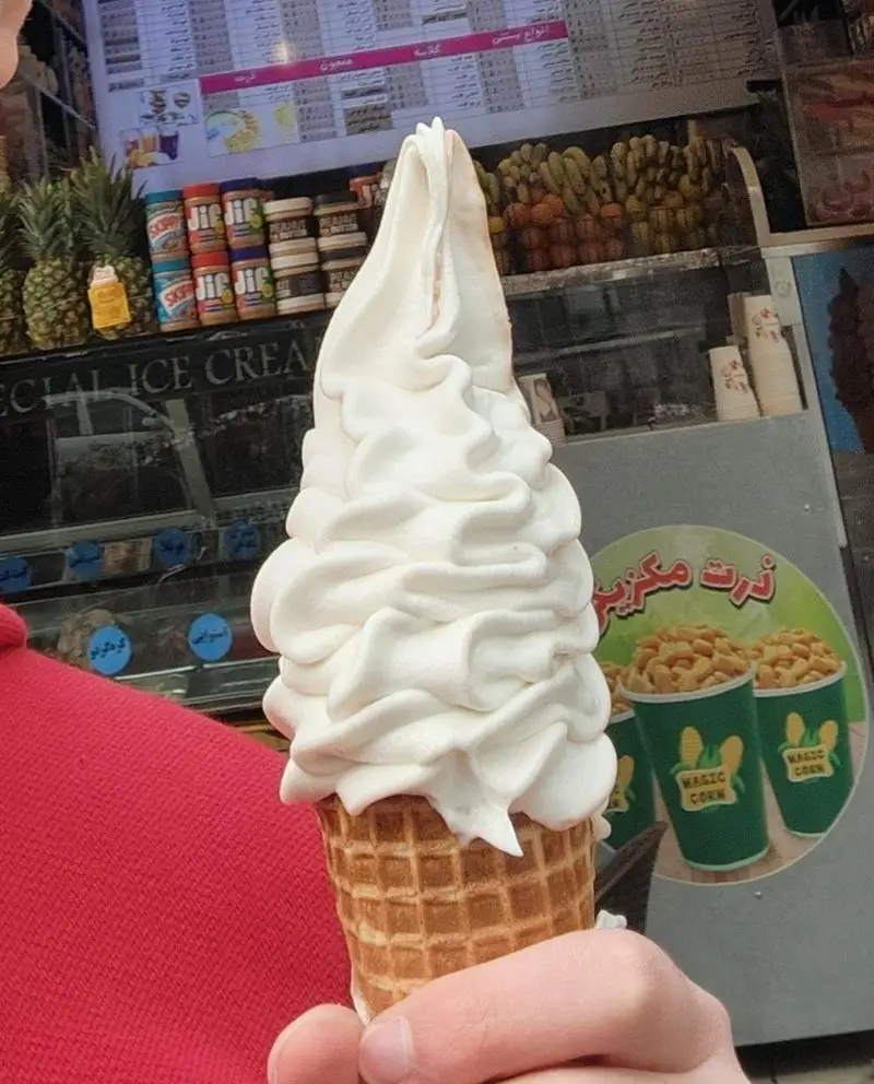 بستنی قیفی محبوب آبمیوه فردین، همراه با میوه‌های تازه مانند آناناس و موز، کره بادام‌زمینی و ذرت مکزیکی در پس‌زمینه.