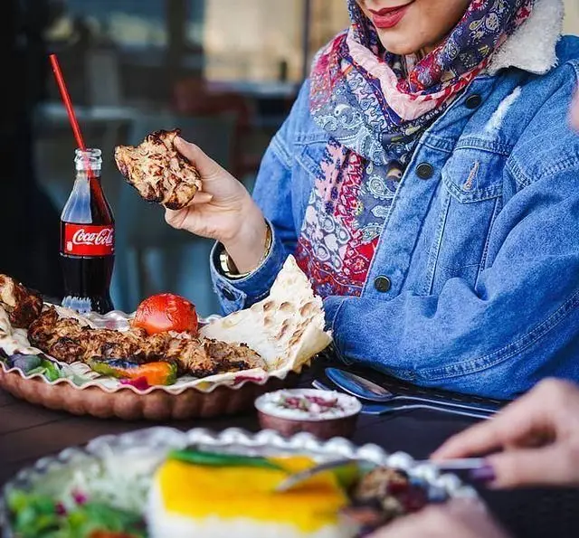 مشتری در رستوران شالِت در حال لذت بردن از کباب ایرانی لذیذ با نان تازه، گوجه کبابی و نوشابه. تجربه ای دلپذیر از غذای سنتی.