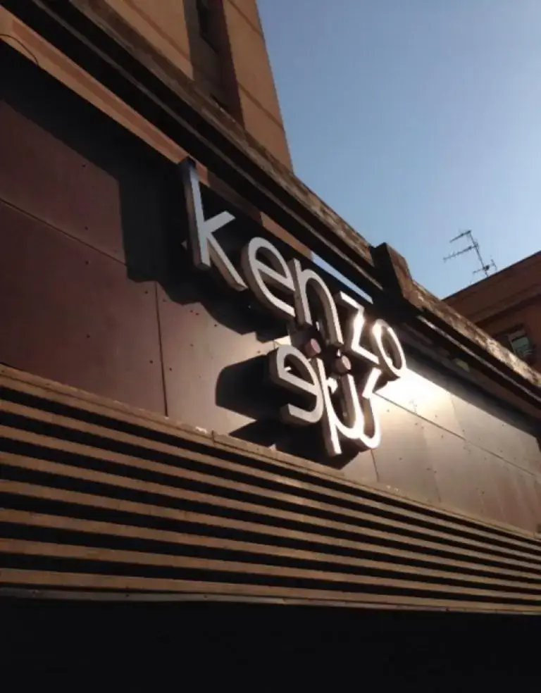 تابلوی برجسته "Kenzo" روی نمای قهوه ای رستوران کنزو، با تابش نور خورشید و آسمان آبی، نمای بیرونی دلنشین این رستوران معروف را به تصویر می کشد.