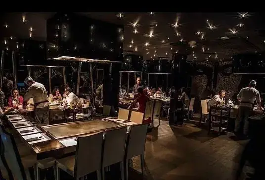 رستوران کنزو: نمای داخلی یک رستوران شیک با نورپردازی ستاره‌ای، مشتریان در حال صرف شام و آشپزان در حال آماده‌سازی غذا