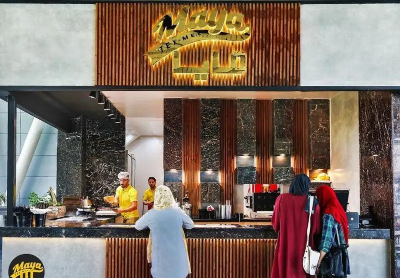 نمایی جذاب از رستوران مايا تِكس مِكس: کارکنان مشغول آماده سازی غذا در پیشخوان شلوغ و مشتریان در حال تماشا و سفارش. دکور مدرن با چوب و سنگ.