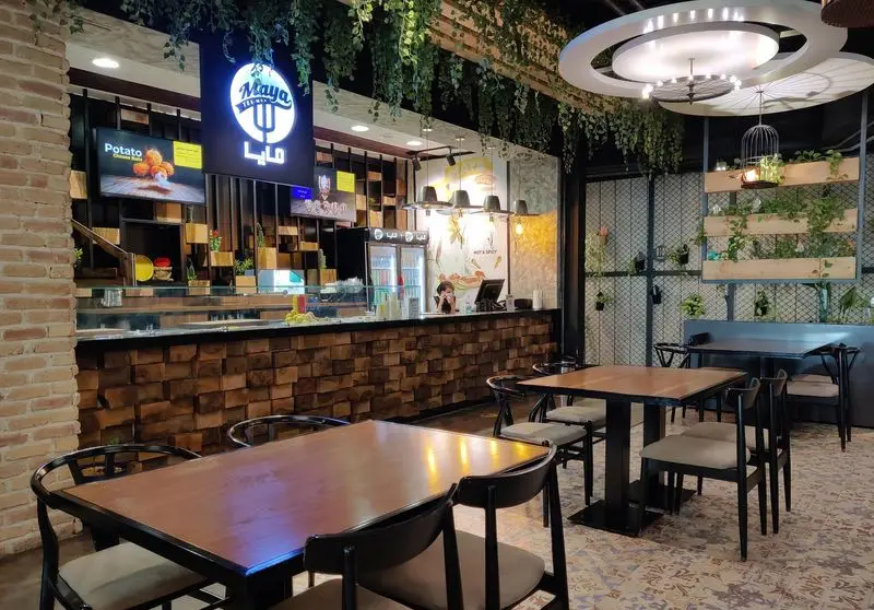 فضای داخلی زیبا و مدرن رستوران مايا تِكس مِكس با کانتر چوبی، تابلو برند روشن، گیاهان دکوری و چیدمان شیک میز و صندلی‌ها.