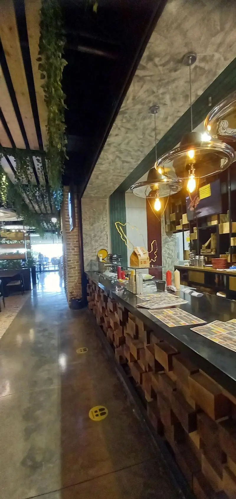 فضای داخلی مدرن و جذاب رستوران مايا تِكس مِكس، شامل کانتر چوبی، نورپردازی خاص، سقف با گیاهان آویز و رعایت فاصله گذاری اجتماعی.