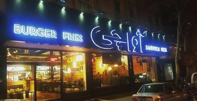 نمای بیرونی رستوران فست فود آواچی در شب با تابلوی نئون درخشان و غذاهایی مانند برگر، سیب زمینی، ساندویچ و پیتزا. محیطی جذاب برای مشتریان.