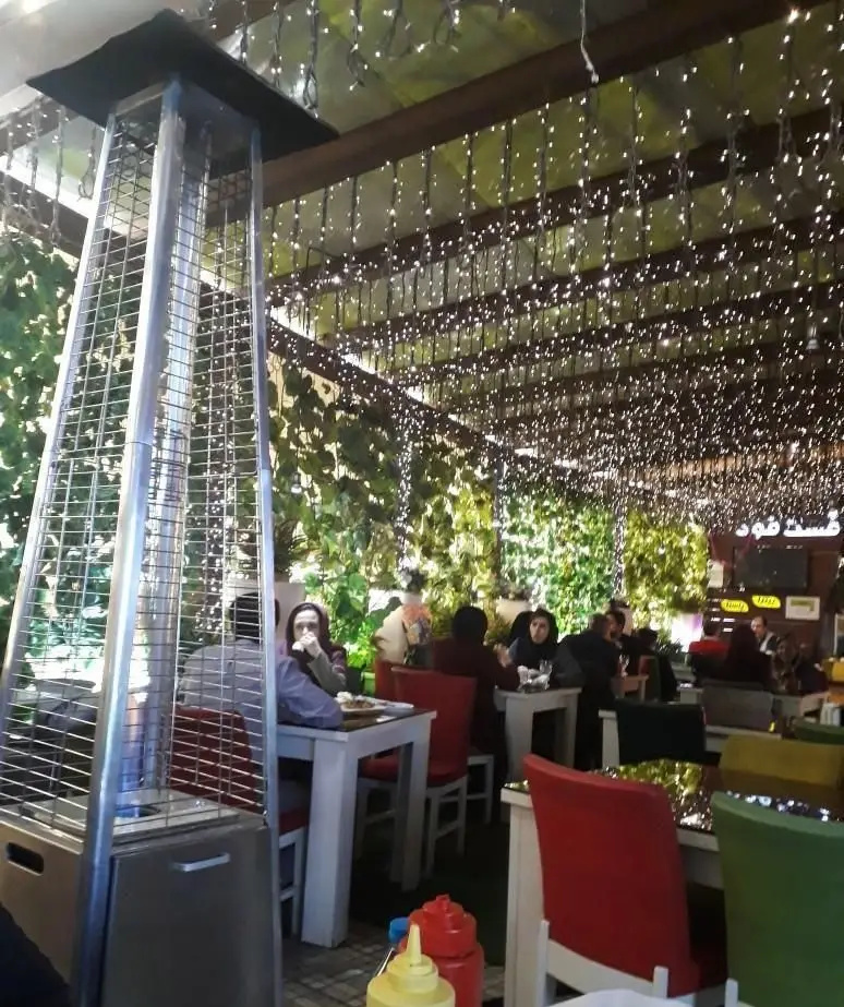 فضای دلنشین و سرسبز فست فود شنتی با نورپردازی زیبا، بخاری قارچی و مشتریانی که در حال صرف غذا هستند. محیطی دنج و شیک.