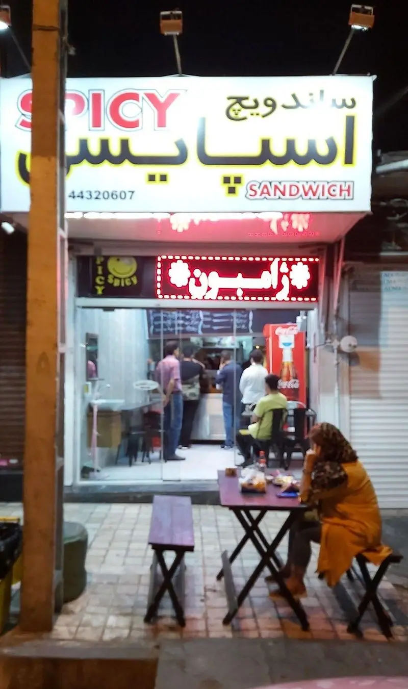 فروشگاه ساندویچ اسپایسی در شب با تابلوهای نئون روشن و میزهای فضای باز. مشتریان در حال خرید و صرف غذای خوشمزه.