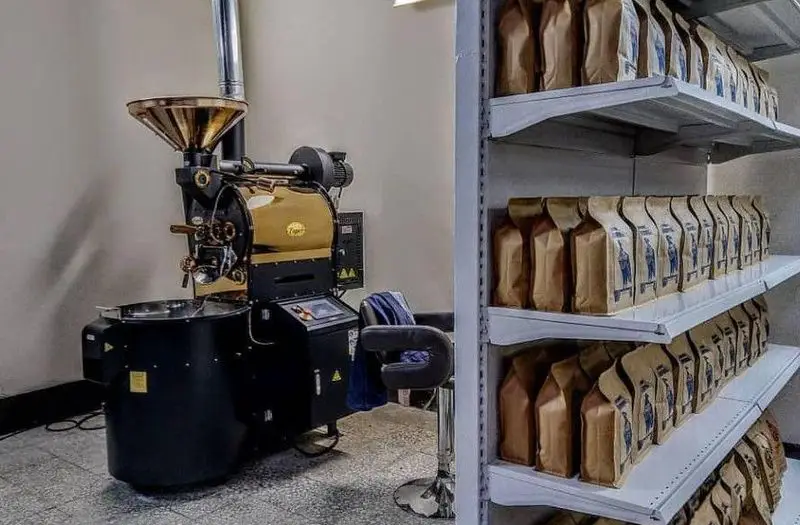 یک دستگاه رست قهوه حرفه‌ای و مدرن در کافه چکاد، با رنگ‌بندی جذاب مشکی و طلایی، به همراه قفسه‌هایی پر از بسته‌های قهوه تازه رست شده برای مشتریان.