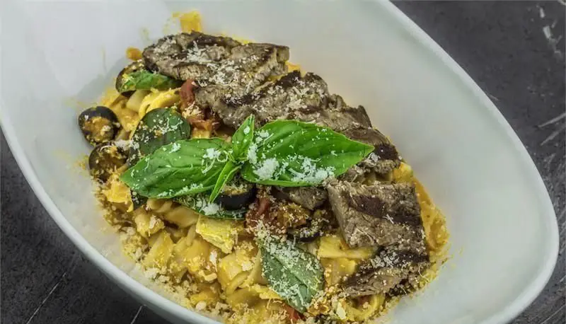 تصویری از پاستای لذیذ با برش‌های استیک آبدار، سس خامه‌ای، زیتون، پنیر پارمزان و ریحان تازه در فودکورت رویال تراس.