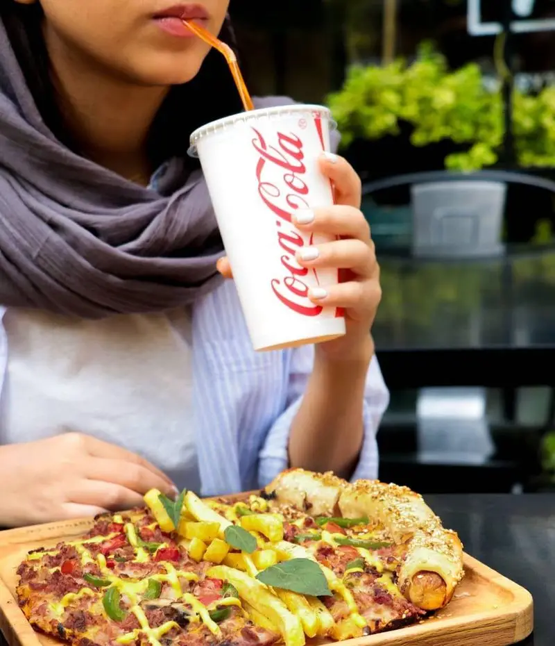 خانمی در حال لذت بردن از پیتزای ویژه فست فود شیلا با سیب زمینی سرخ کرده، قارچ، و سوسیس در لبه، همراه با نوشابه کوکا کولا.
