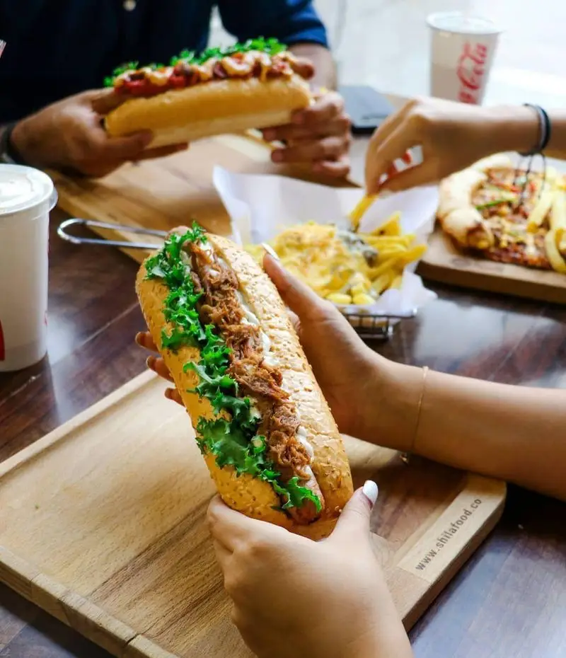 دو نفر در فست فود شیلا در حال لذت بردن از هات داگ های خوشمزه، همراه با پیتزا و سیب زمینی سرخ کرده در میزی پر از غذا.