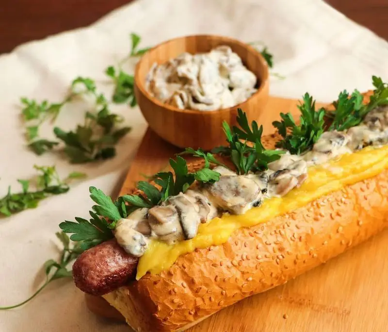 ساندویچ هات داگ سوسیس با سس قارچ و پنیر آب شده، تزئین شده با جعفری تازه از فست فود شیلا، یک انتخاب لذیذ و پرطرفدار.