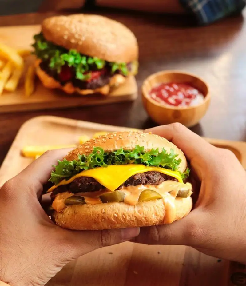 تصویر نزدیک از چیزبرگر لذیذ در دست مشتری، با سیب زمینی و سس قرمز در پس‌زمینه. تجربه غذایی عالی در فست فود شیلا.