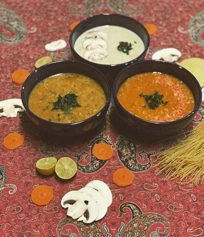 سفره‌ای از انواع سوپ‌های خانگی بی بی: سوپ قارچ، سبزیجات و عدس قرمز، تازه و گرم با تزیینات هویج، لیمو و قارچ تازه.