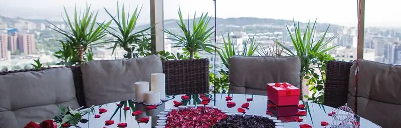 میز رمانتیک و زیبا در فضای باز رستوران ارکیده اقدسیه، با گلبرگ‌های رز، شمع و هدیه، ایده‌آل برای یک قرار عاشقانه با منظره شهر.