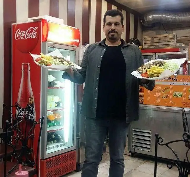 مردی در شهر پیتزا با افتخار دو بشقاب کباب و سیب زمینی سرخ کرده را در مقابل یخچال نوشابه و تابلوی منوی رستوران به نمایش می گذارد.