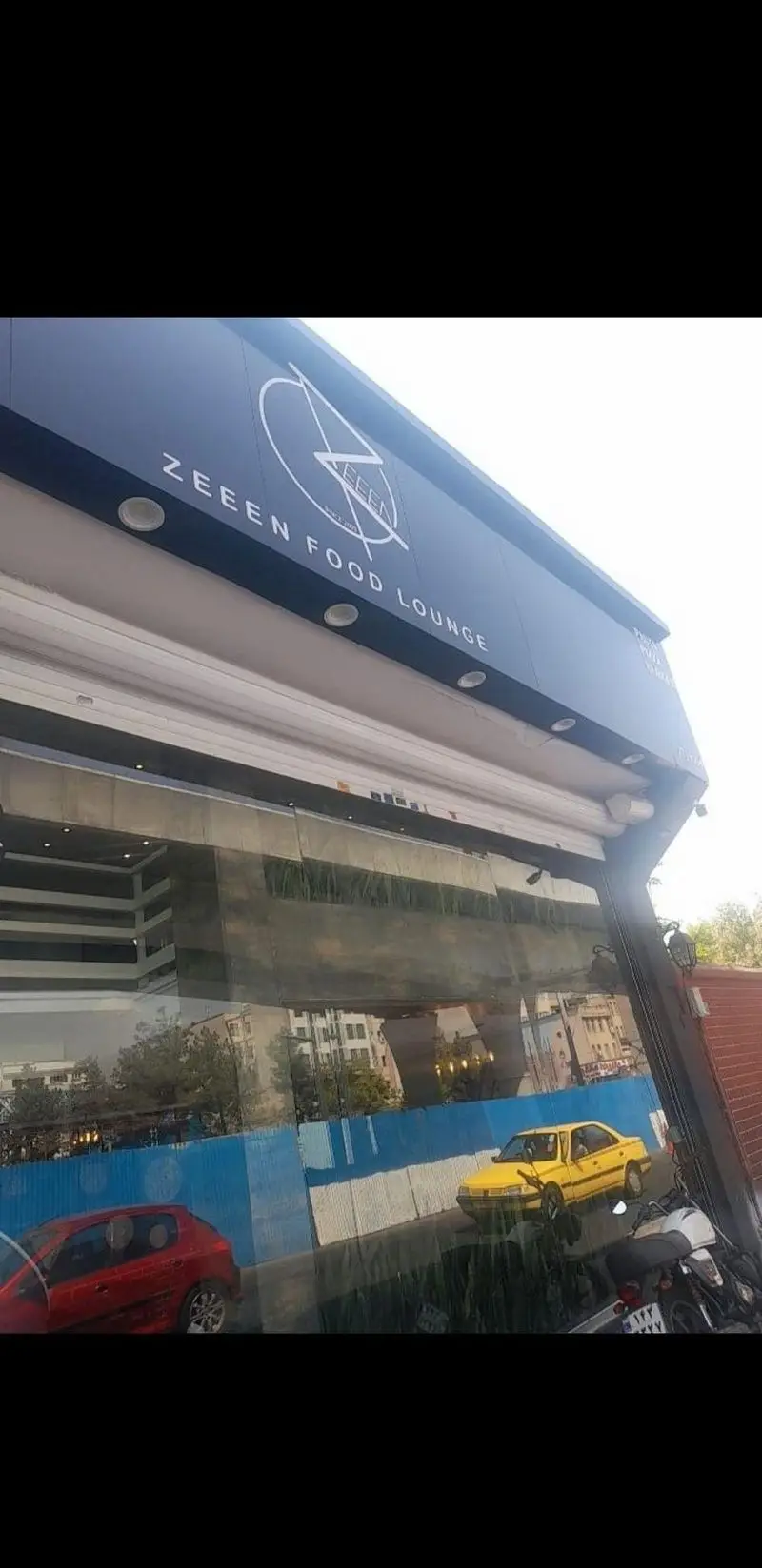 تصویری از نمای خارجی و تابلوی شیک فست فود زین (ZEEEN FOOD LOUNGE) با بازتاب محیط شهری و خودروها در شیشه‌ها، مکانی مدرن برای غذای دلپذیر.