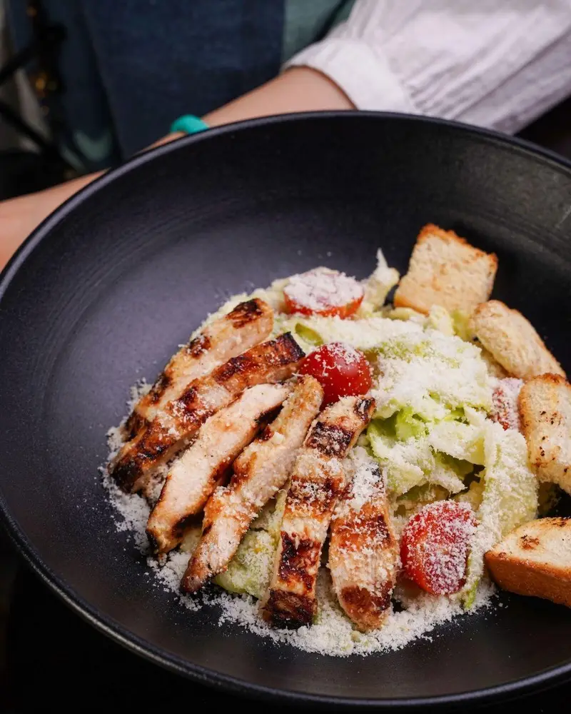 لذت چشیدن سالاد سزار با تکه‌های مرغ گریل ترد و خوش‌طعم، کاهو تازه، گوجه گیلاسی و پنیر پارمزان در کافه رستوران عجمان.