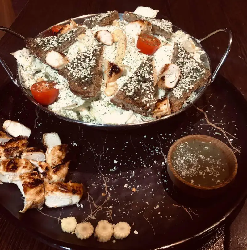سالاد سزار لوکس کافه رستوران عجمان با مرغ گریل آبدار، نان تست کنجدی و گوجه فرنگی، به همراه سس مخصوص در ظرف شیک و بزرگ.