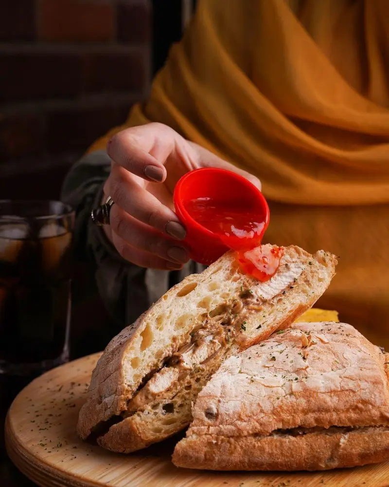 ساندویچ چیکن خوشمزه کافه رستوران عجمان با نان چاباتا و سس قرمز. تجربه‌ی غذایی دلپذیر در فضایی دنج و عالی.