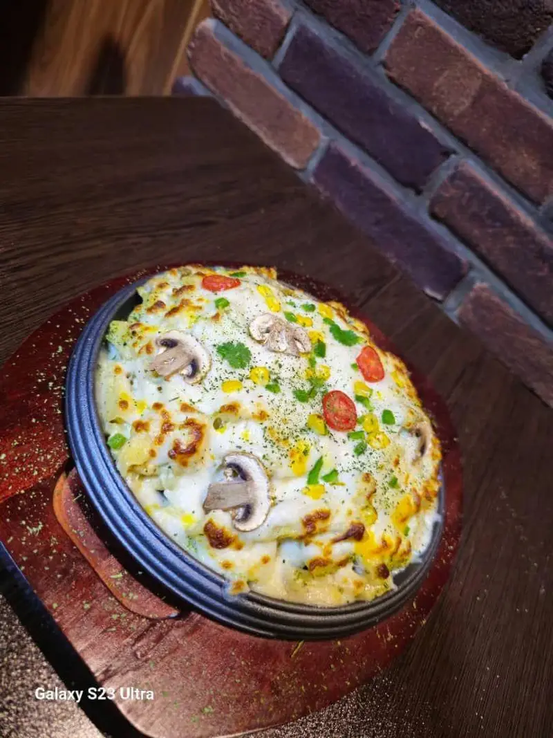 تصویری جذاب از یک گراتن پنیری داغ و لذیذ با قارچ، ذرت و گوجه فرنگی، سرو شده بر روی میز چوبی کافه رستوران عجمان.