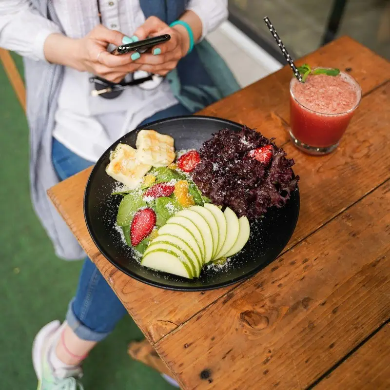 مشتری در کافه رستوران عجمان مشغول چک کردن موبایل کنار سالاد میوه و سبزیجات سالم با پنیر و اسموتی خنک، تجربه‌ای دلپذیر.