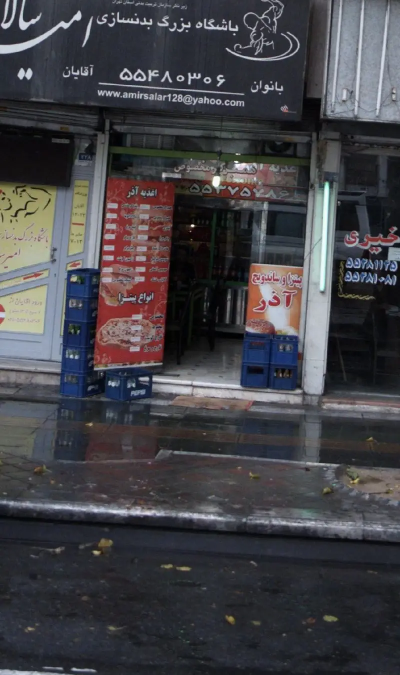 نمای خارجی مغازه پیتزا و ساندویچ آذر، یک فست فود محبوب با منوی متنوع از انواع پیتزا و ساندویچ‌های تازه در یک روز بارانی.