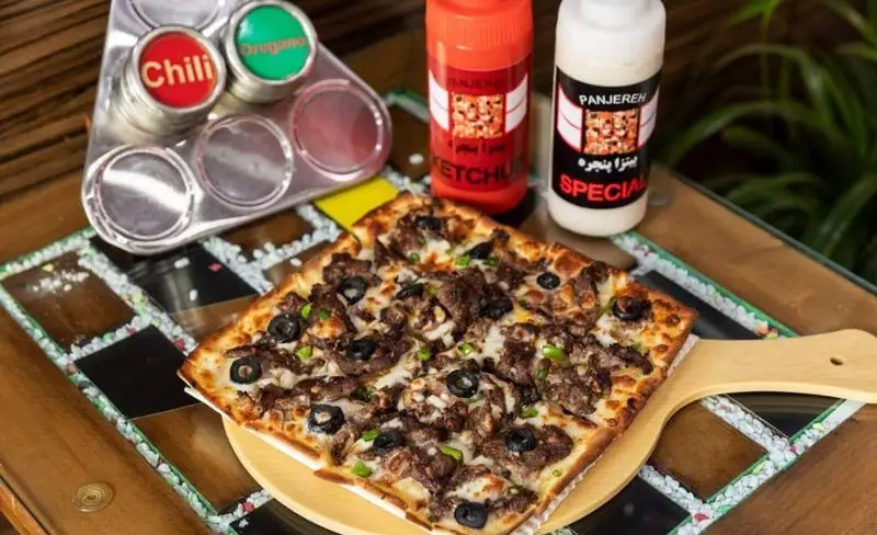 پیتزای گوشت و زیتون چهارگوش از پیتزا پنجره، همراه با ادویه چیلی و آویشن، سس کچاپ و سس مخصوص پانجره روی میزی زیبا.