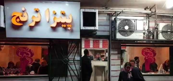 پیتزا ترنج: نمای بیرونی شلوغ فست فود پیتزا ترنج در شب با تابلوی روشن و مشتریان در حال صرف غذا.